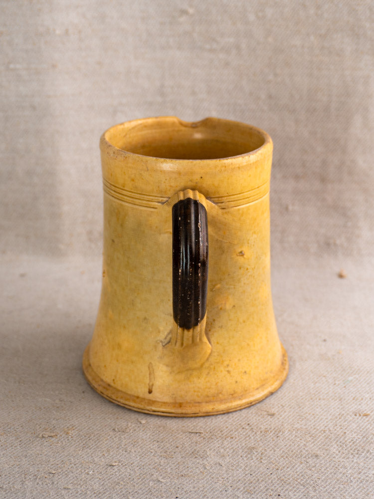 Antique French Pichet Jaune, yellow glazed earthenware mug with dark brown handle on beige textured surface.