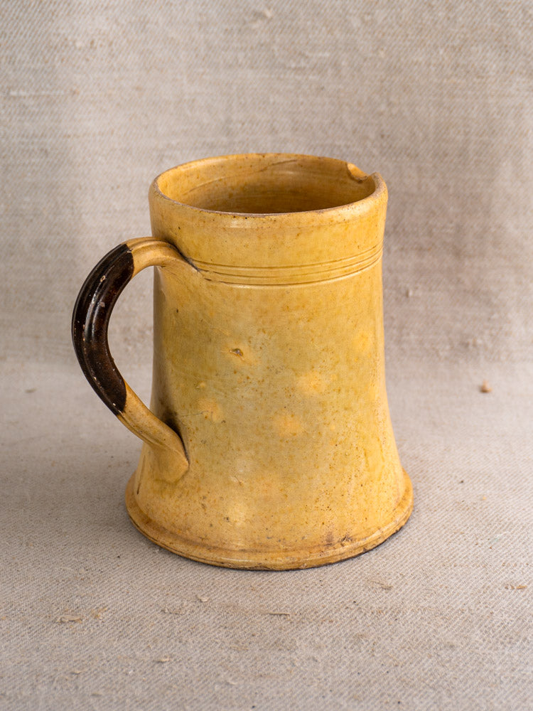 Antique French yellow glazed earthenware pichet with dark brown handle, rustic finish on textured beige fabric background.