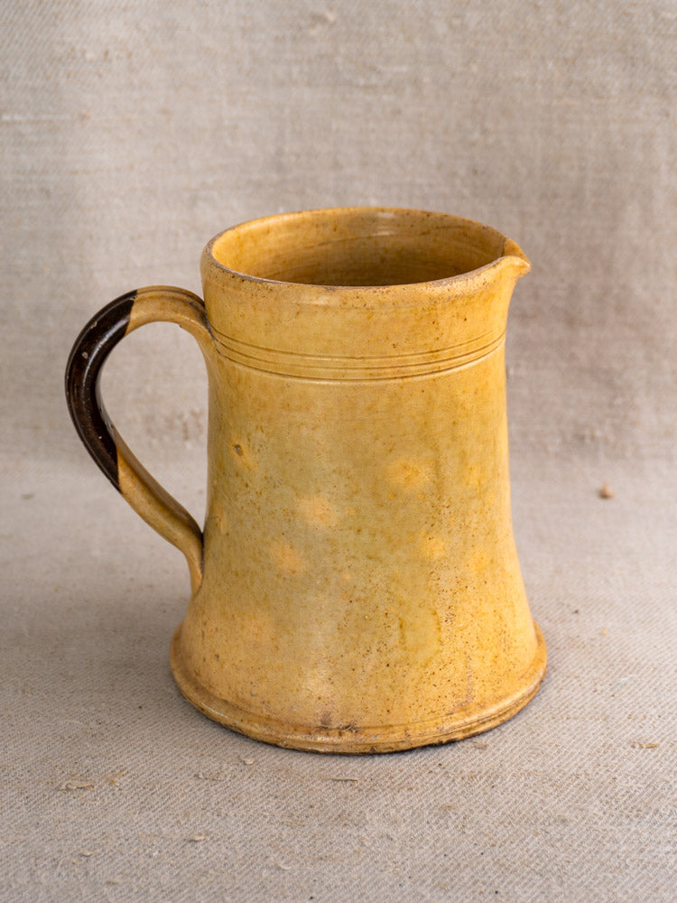 Antique French Pichet Jaune yellow earthenware pitcher with wide base, narrow top, and dark brown handle on beige fabric.