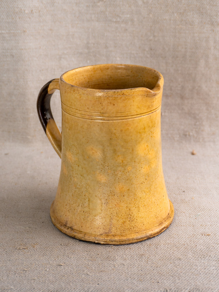 Antique French yellow glazed earthenware pitcher with flared base, curved handle, and small spout on beige fabric background.