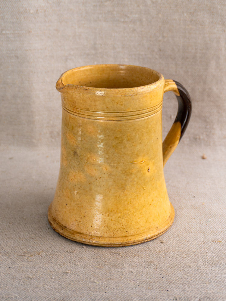 Antique French yellow glazed earthenware pichet with dark brown handle, shown on textured beige fabric.