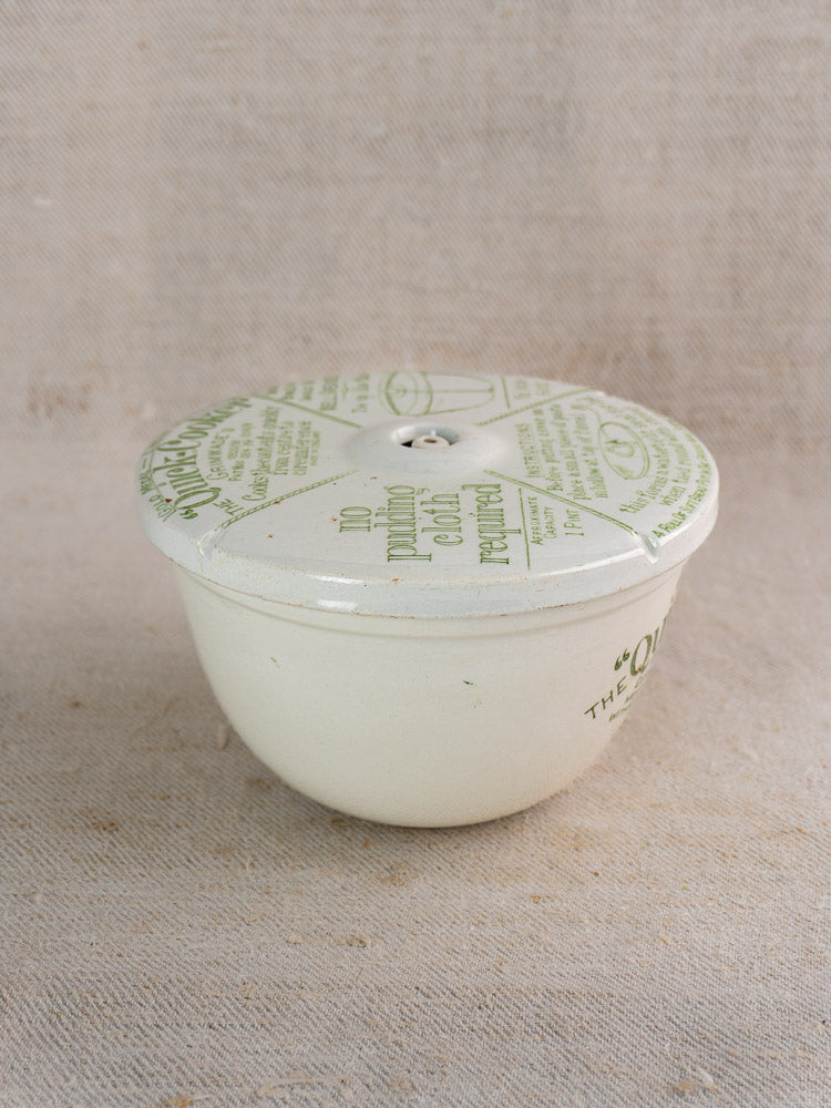 Antique Grimwade Quick Cooker pudding steamer, c.1911, white ceramic with green script, on beige textured fabric.