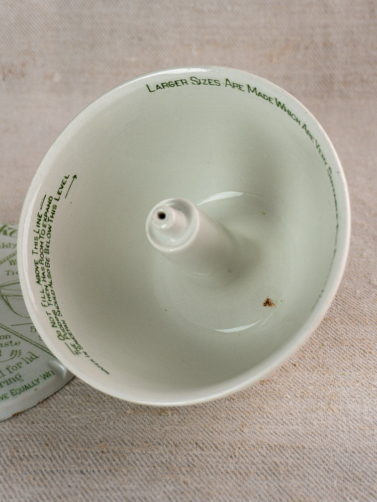 Antique Grimwade English pudding steamer, white ceramic with green rim text and central spout, on beige textured surface.
