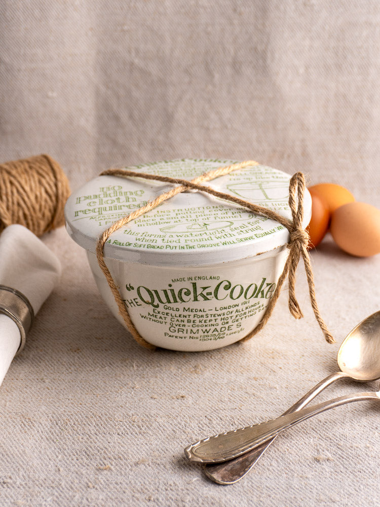 Antique English Grimwade Quick Cooker pudding steamer with fitted lid and green label on textured beige fabric.