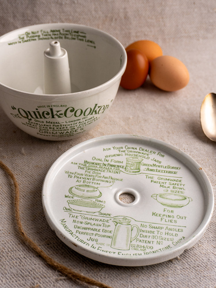Antique English pudding steamer with green script, ceramic lid, and illustrated bowl beside eggs and spoon on beige surface.