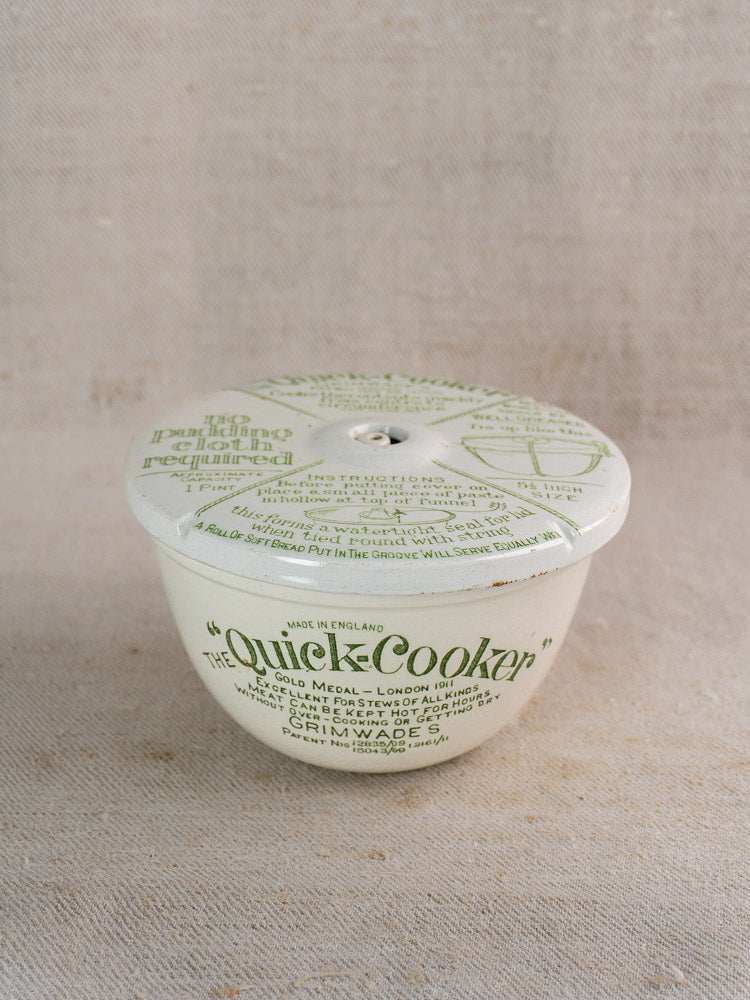 Antique English Grimwade Quick Cooker pudding steamer with instruction-printed lid on beige textured fabric.