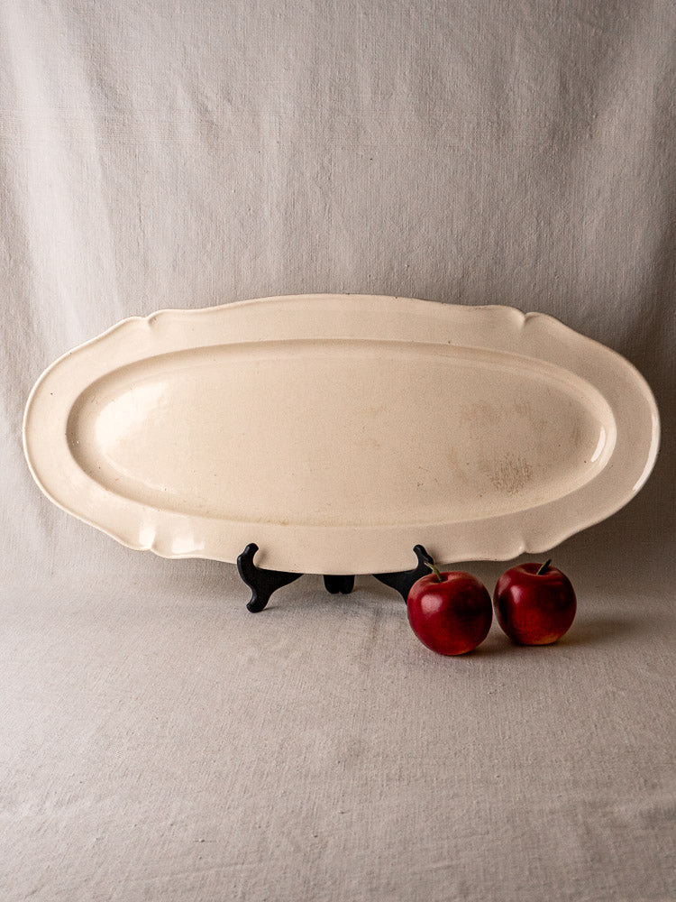 Antique French faience meat platter, cream earthenware, on black stand with red apples, fabric backdrop.