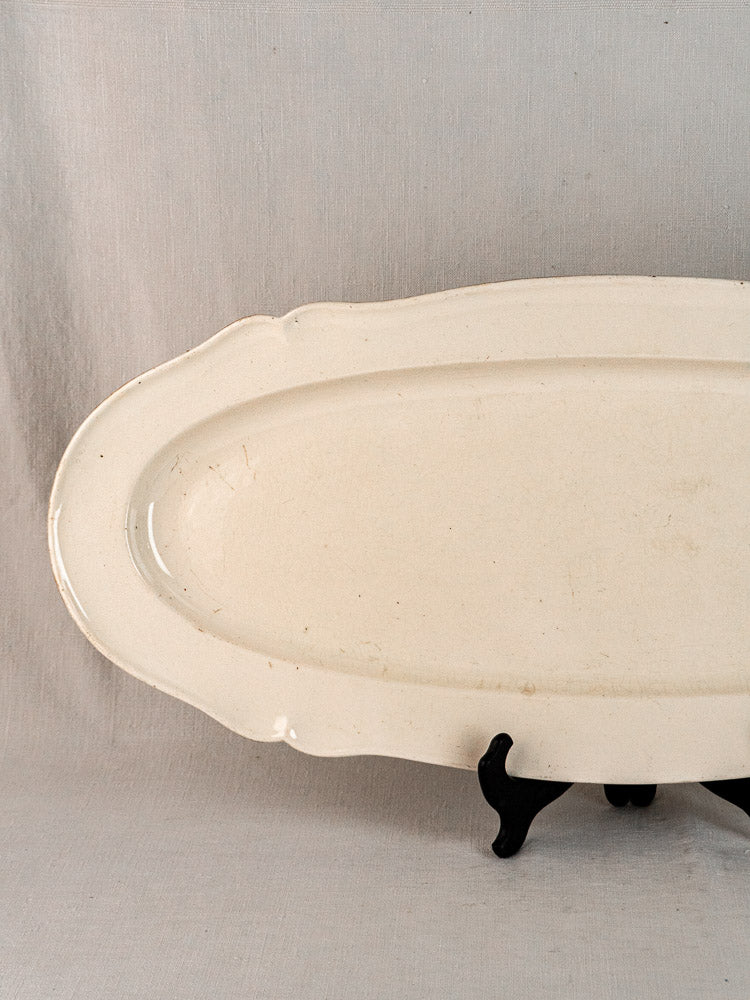 Oval antique French faience meat platter with wavy rim on black stand, neutral fabric backdrop.