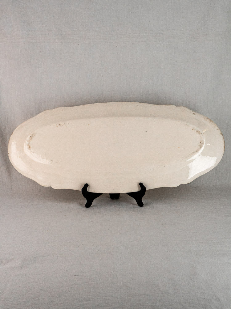 Antique French faience oval meat platter with scalloped edges on black stand, gray background.