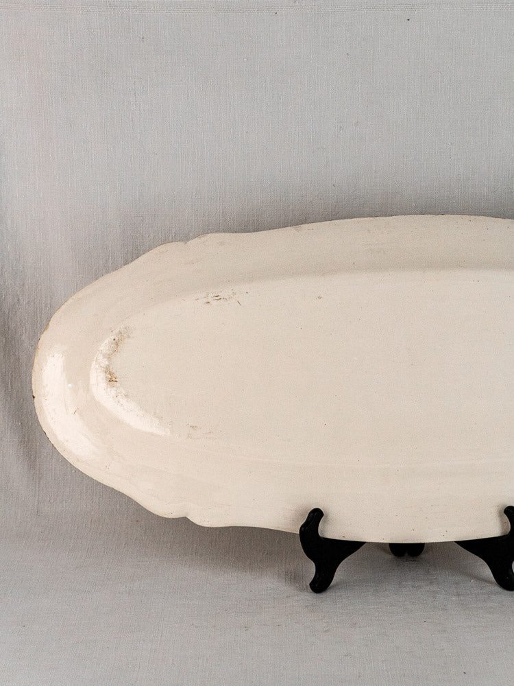 Antique French oval white ceramic meat platter with scalloped edges on black stand, gray fabric backdrop.