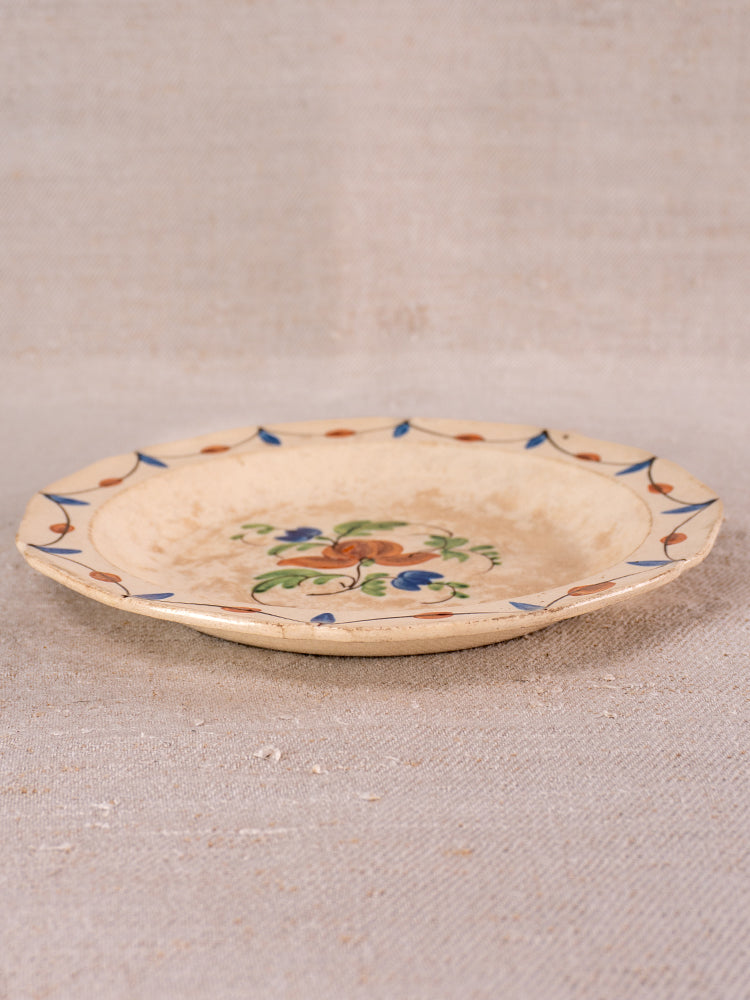 Antique French faience plate with hand-painted Normandy floral motif, blue and brown border on neutral fabric background.