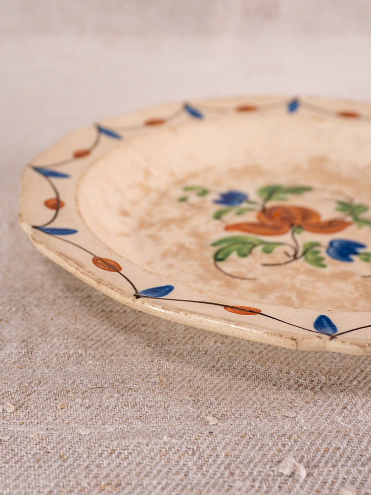 Antique French faience plate with central orange and green floral motif, blue-orange leaf border, on textured beige surface.