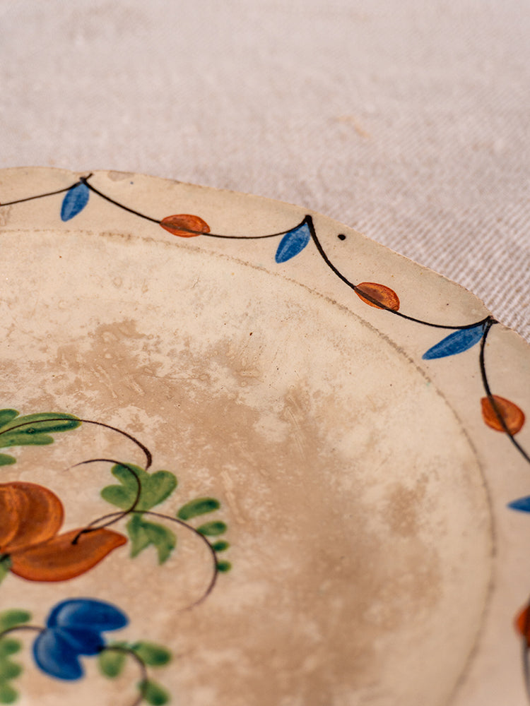Antique French faience plate with hand-painted blue, orange, and green Normandy floral motifs on a cream ground and worn edges.