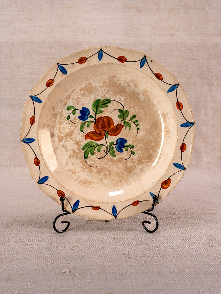 Antique French faience plate, Normandy, early 19th century, scalloped edge, floral motif, on metal stand with neutral fabric backdrop.