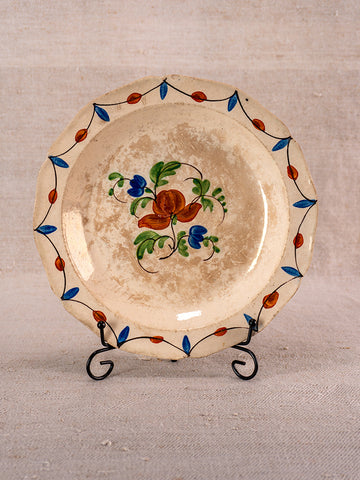 Antique French faience plate, Normandy, early 19th century, scalloped edge, floral motif, on metal stand with neutral fabric backdrop.