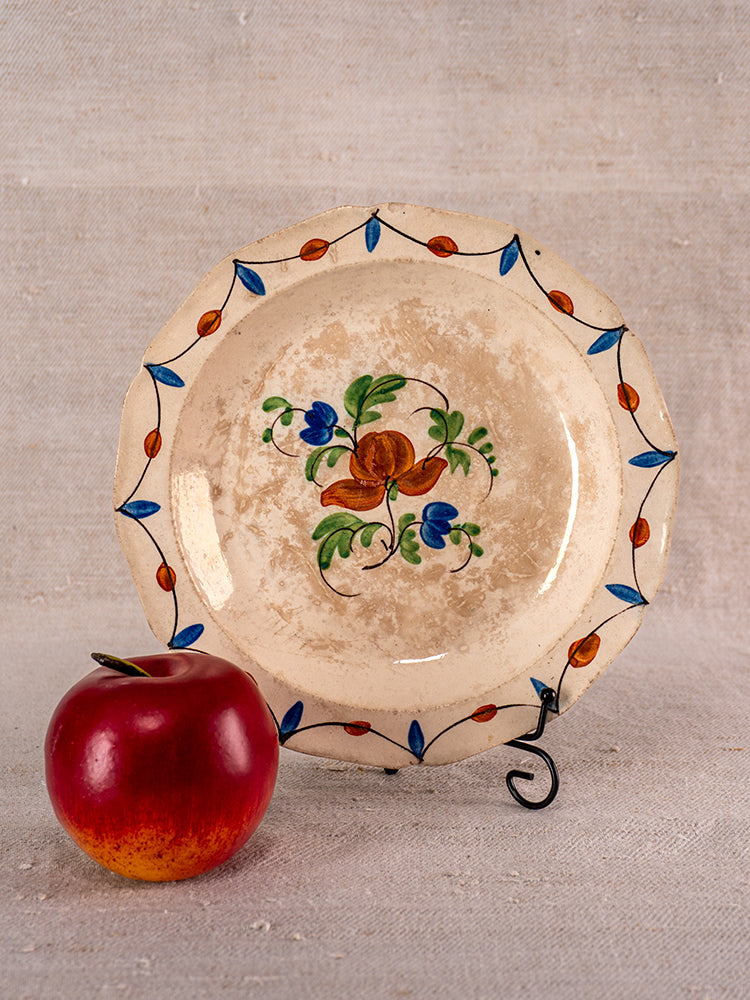 Antique French faience plate with Normandy floral design on metal stand, red apple in front, beige textured background.