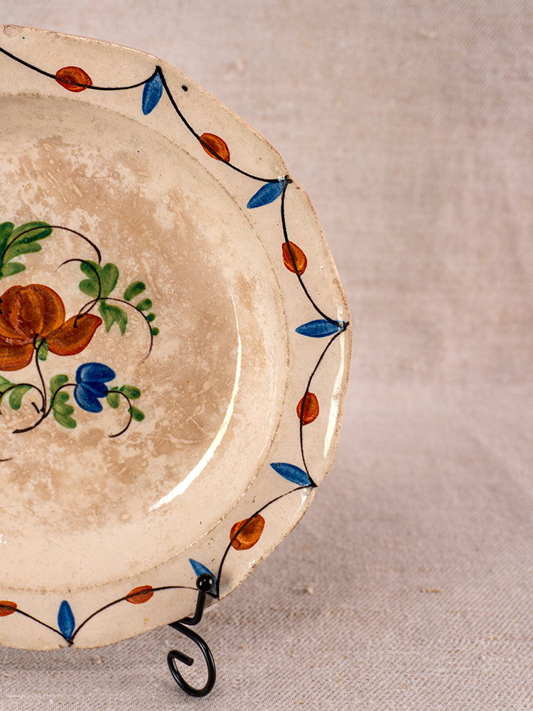 Antique French faience plate with vivid orange, green, and blue florals on black stand against beige backdrop.