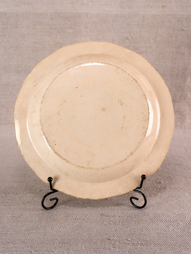 Antique French faience plate with off-white glaze and irregular rim on black metal stand, beige textured fabric backdrop.