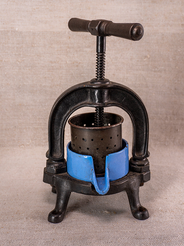 Antique French cast iron fruit press with blue enamel base and perforated metal cylinder on neutral fabric.