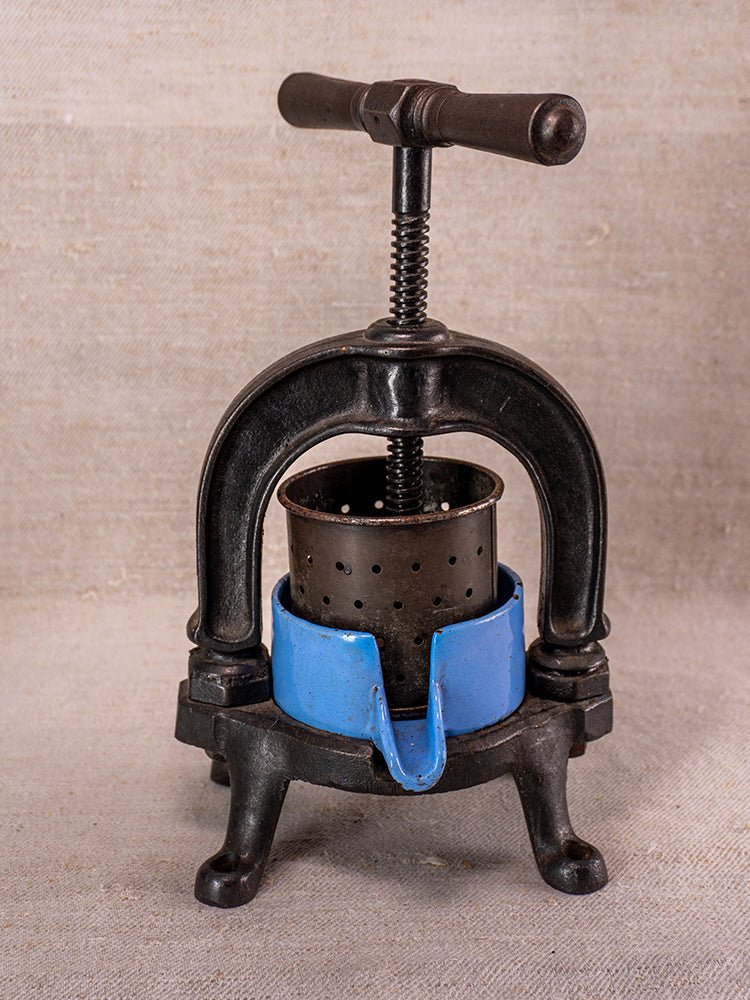Antique French cast iron fruit press with blue enamel base and perforated metal cylinder on neutral fabric.