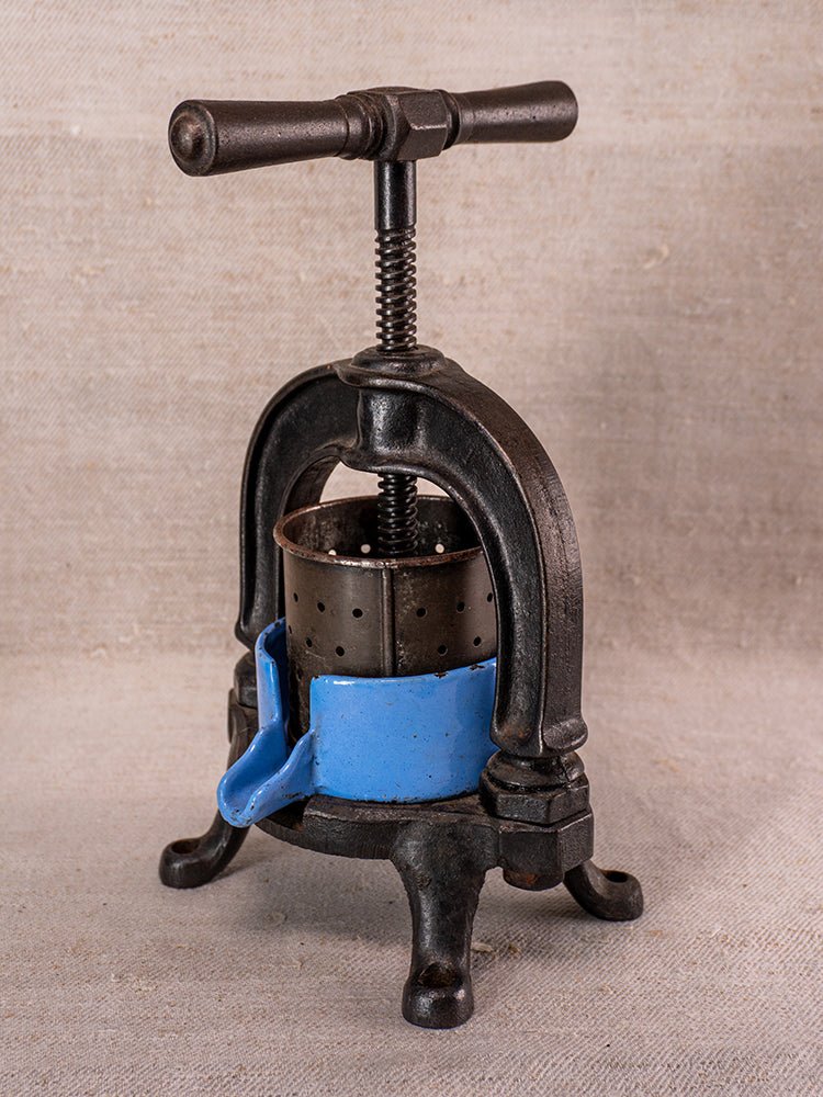 Antique French fruit press in cast iron with blue enamel band and handle, on neutral fabric background.