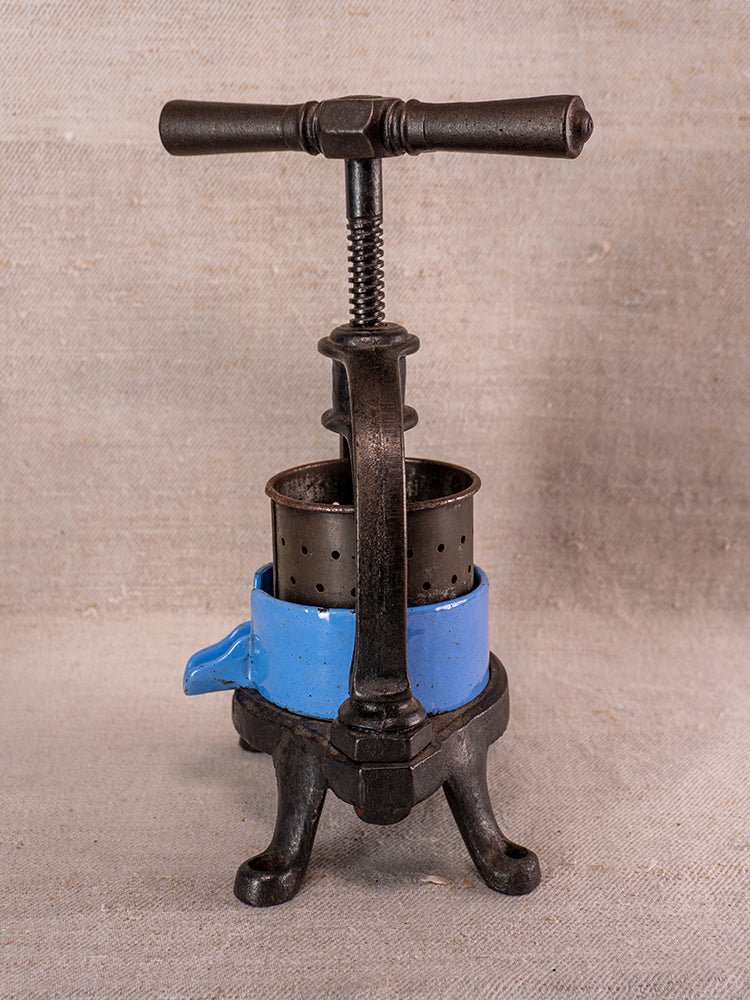 Antique French cast iron fruit press with blue enamel, crank handle, and perforated basket on fabric background.