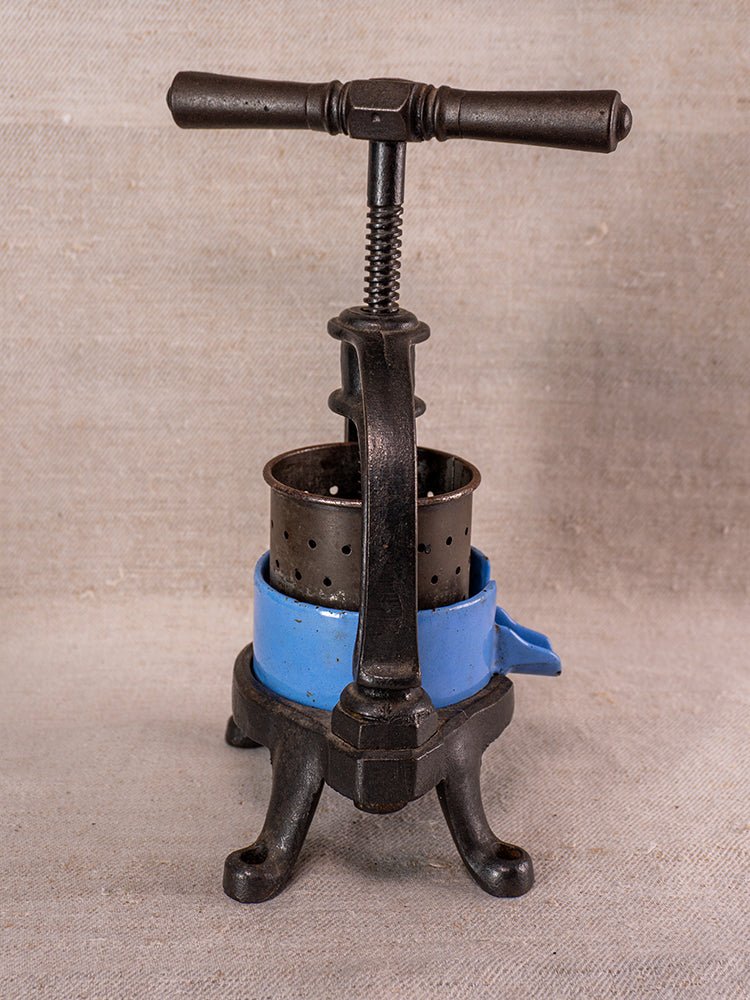 Antique French cast iron fruit press with blue enamel ring and black screw handle on fabric backdrop.