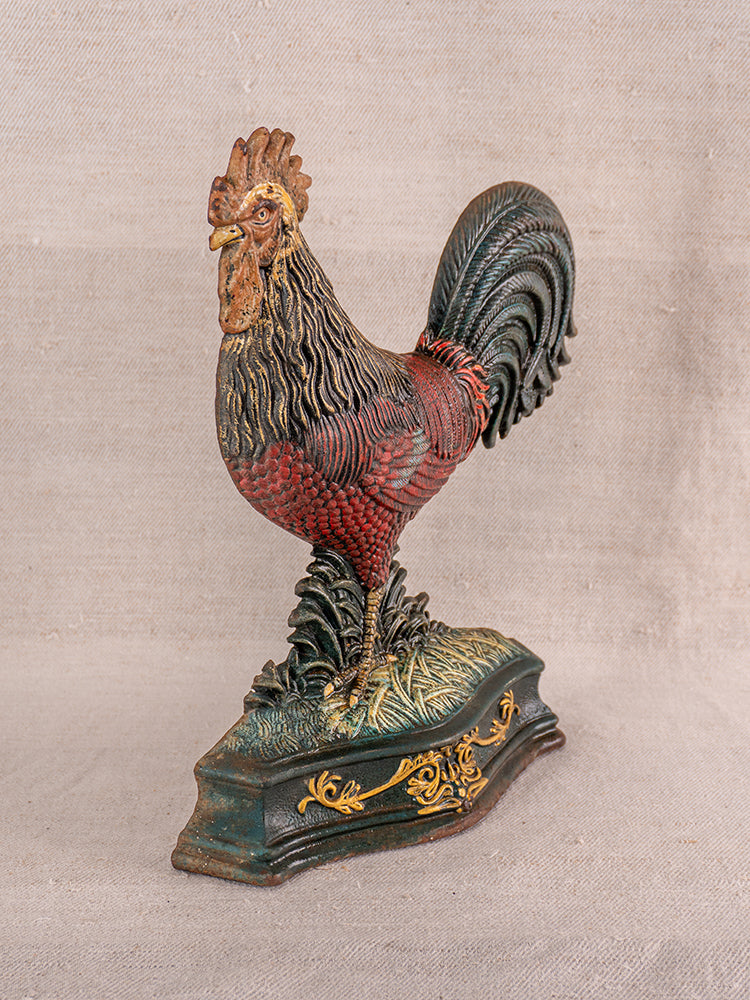 Antique French cast iron rooster doorstop with polychrome red and green feathers, on decorative base, set on neutral fabric.