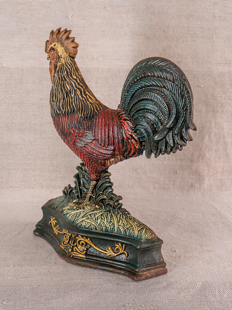 Antique French cast iron rooster doorstop with red, green, and gold polychrome on ornate base, on textured beige fabric.