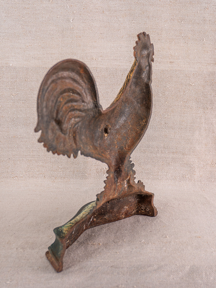 Antique French cast iron rooster doorstop with polychrome finish, curved base, on textured beige fabric.