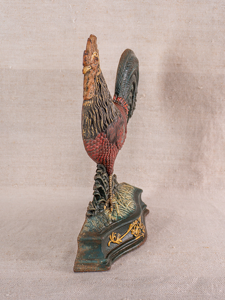 Late 19th-century French cast iron rooster doorstop with polychrome finish on a green and gold base, beige fabric backdrop.