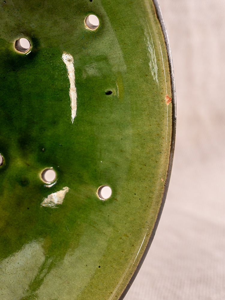 Antique French berry drainer set in green glazed earthenware with round holes and slight edge chip, on neutral textured background.