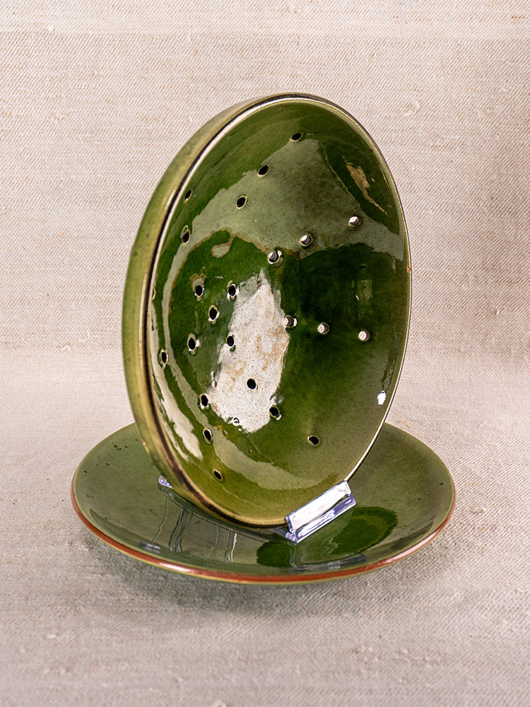 Antique French green glazed earthenware berry drainer set, pierced and solid plates on stand, neutral fabric backdrop, mid 20th century.