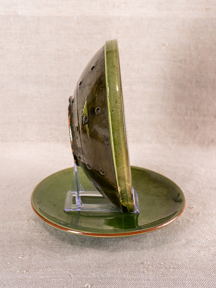 Antique French green glazed earthenware berry drainer set on clear stand, neutral fabric background.