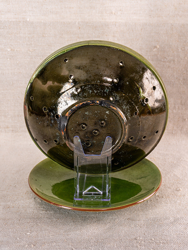 Antique French green glazed earthenware berry drainer set with bowl, clear stand, and matching plate on beige background.