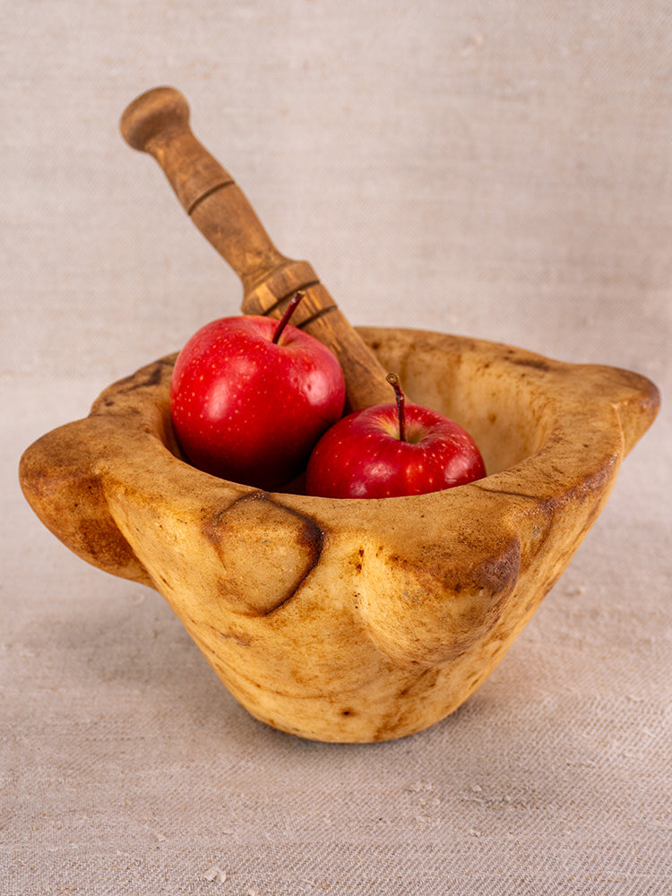 Antique French marble mortar and pestle with classic Provencal lines, two red apples inside, on a neutral fabric background.