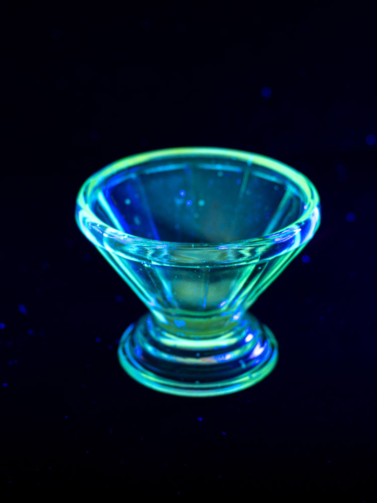 Set of 12 antique French Art Deco uranium glass sherbet coupes glowing green and blue under black light, dark backdrop.
