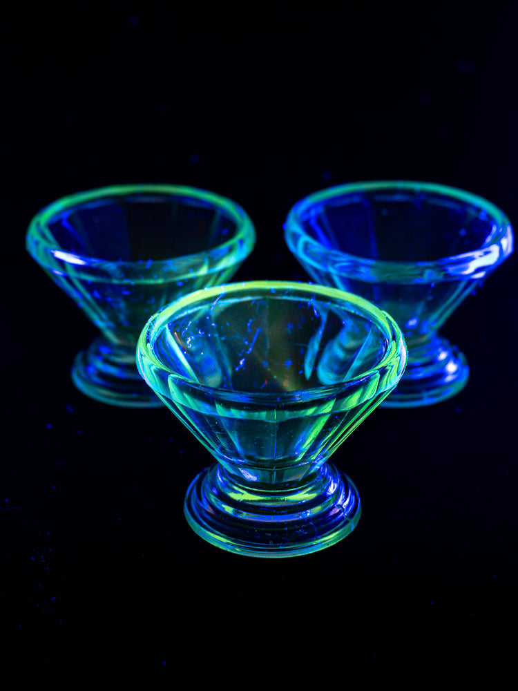 Set of 12 antique French uranium glass sherbet coupes glowing green and blue under blacklight, Art Deco curves, faceted design.