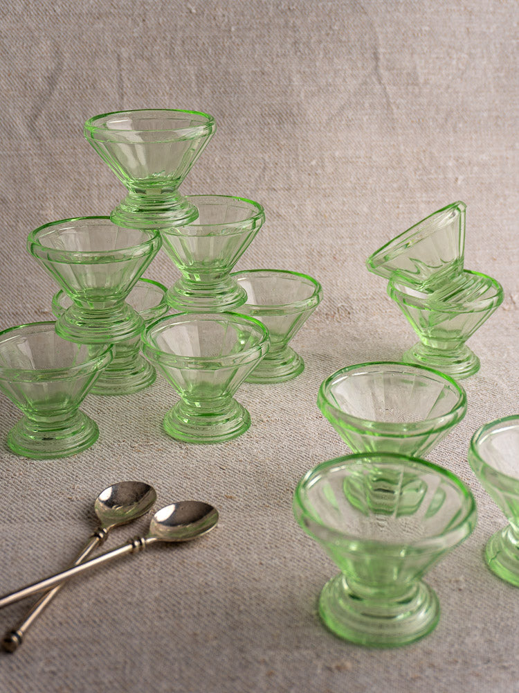 Set of 12 antique French uranium glass sherbet coupes with Art Deco design, stacked with two silver spoons on beige fabric.