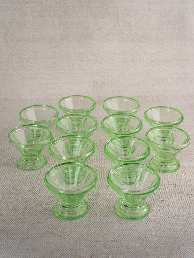 Set of 12 antique French uranium glass sherbet coupes with green Art Deco design, arranged on beige textured surface.