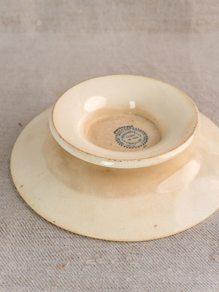 Antique French Boulenger Choisy-le-Roi compote plate upside down on beige fabric, faded maker’s mark and blue base design visible.