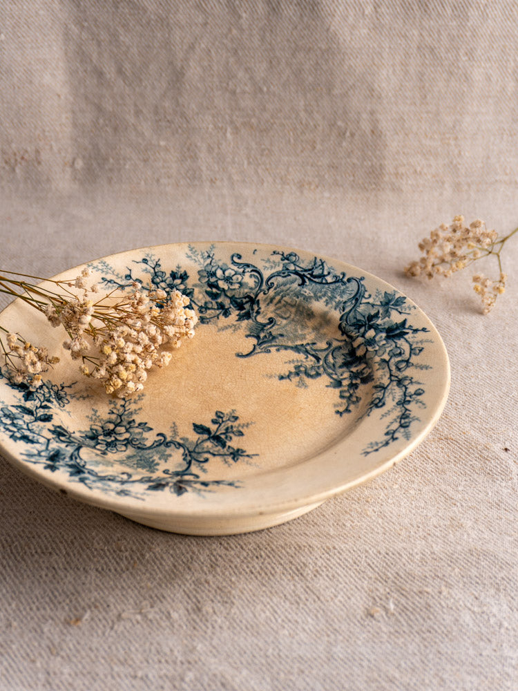 Antique French Boulenger Choisy-le-Roi Pompadour compote plate with blue floral pattern, gentle wear, dried white flowers nearby.