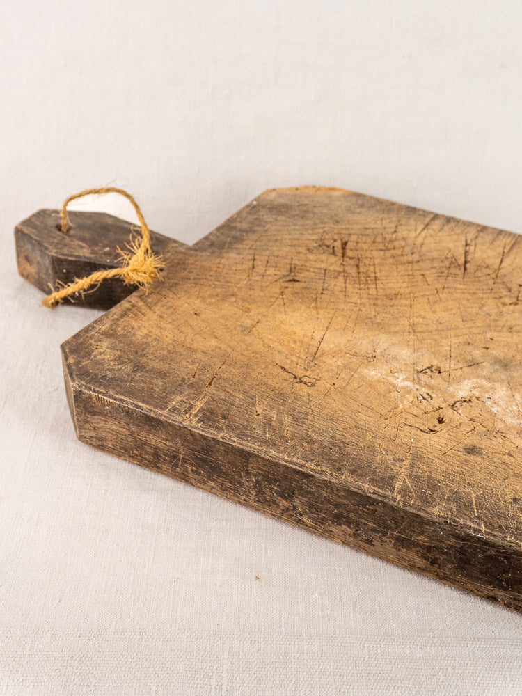 Antique French cutting board with hand-carved diamond handle, visible scratches, and frayed string loop on light fabric background.
