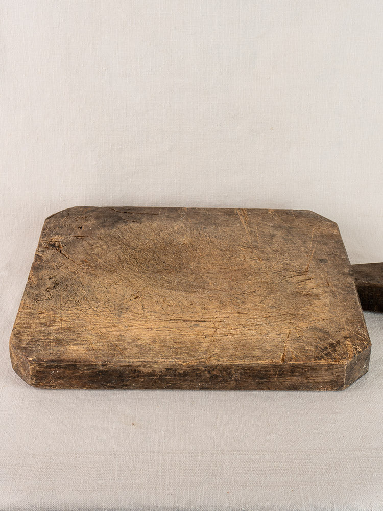 Antique French cutting board with diamond handle, rounded corners, and aged patina on light fabric background.