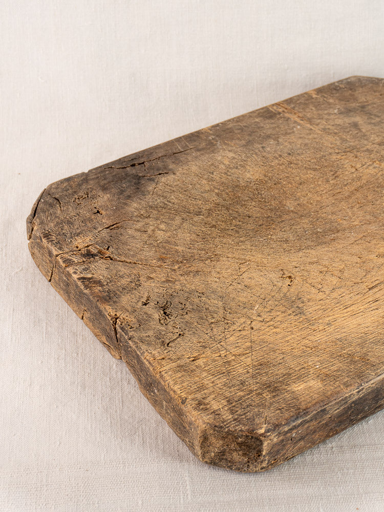 Antique French cutting board, hand carved with diamond handle, rounded edges and cracks, on light fabric surface.