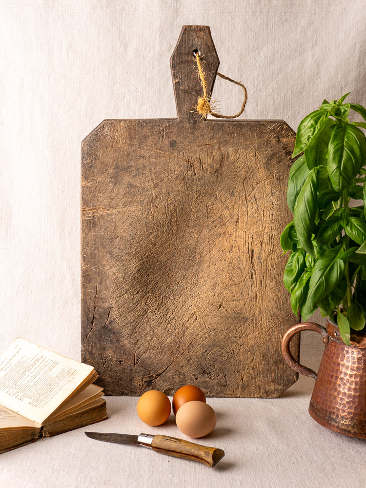 Antique French cutting board with diamond-carved handle, string, eggs, open cookbook, knife, and copper jug with basil.