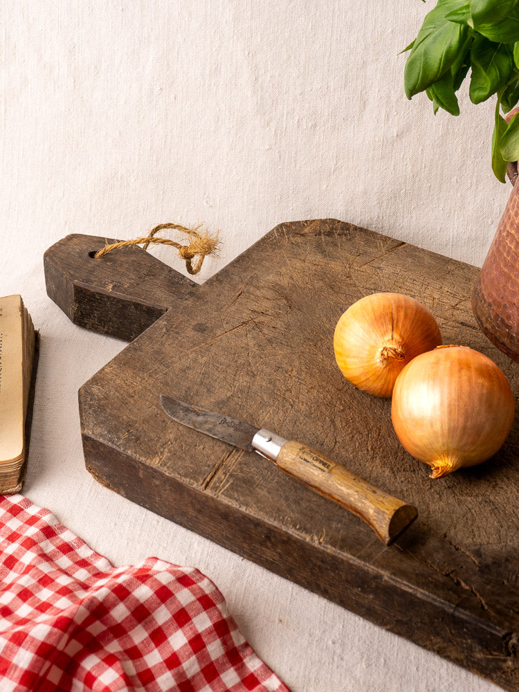 Antique French cutting board with carved diamond handle, displayed with onions, knife, checkered cloth, book, and basil plant.