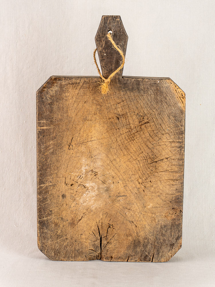 Antique French cutting board circa 1850 with diamond-carved handle, rectangular form, worn wood, rounded corners, and frayed rope.