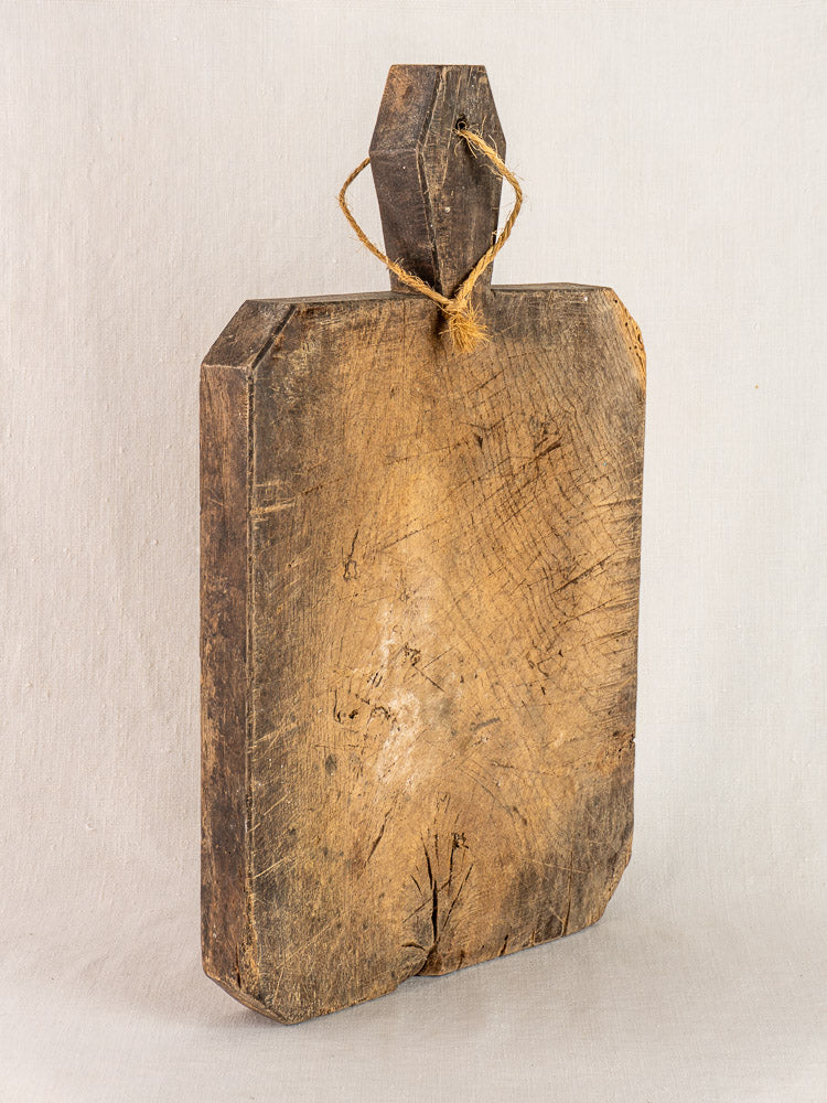 Antique French cutting board circa 1850 with hand-carved diamond handle and twine, upright against a light background.