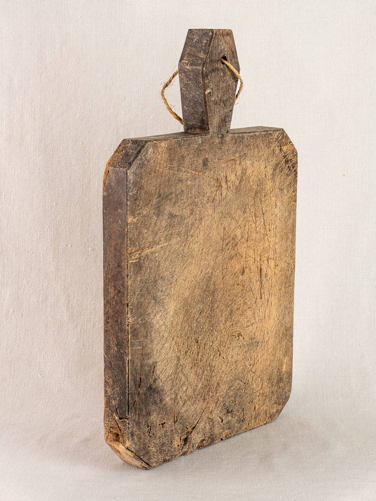 Antique French cutting board, circa 1850, with hand-carved diamond handle and rope, upright on light background.